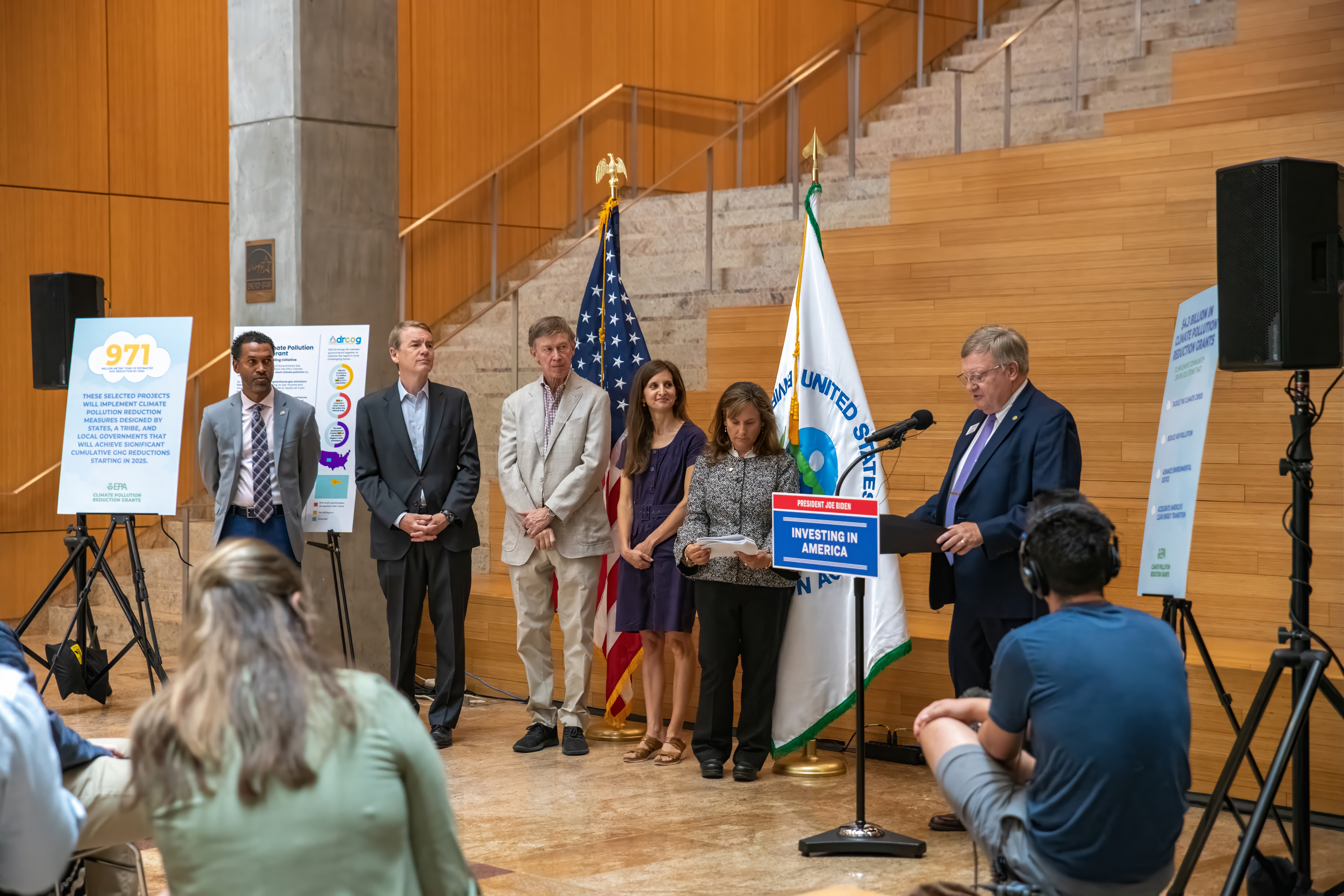 Five people stand in front of flags stand to the left of a man speaking at a lectern.