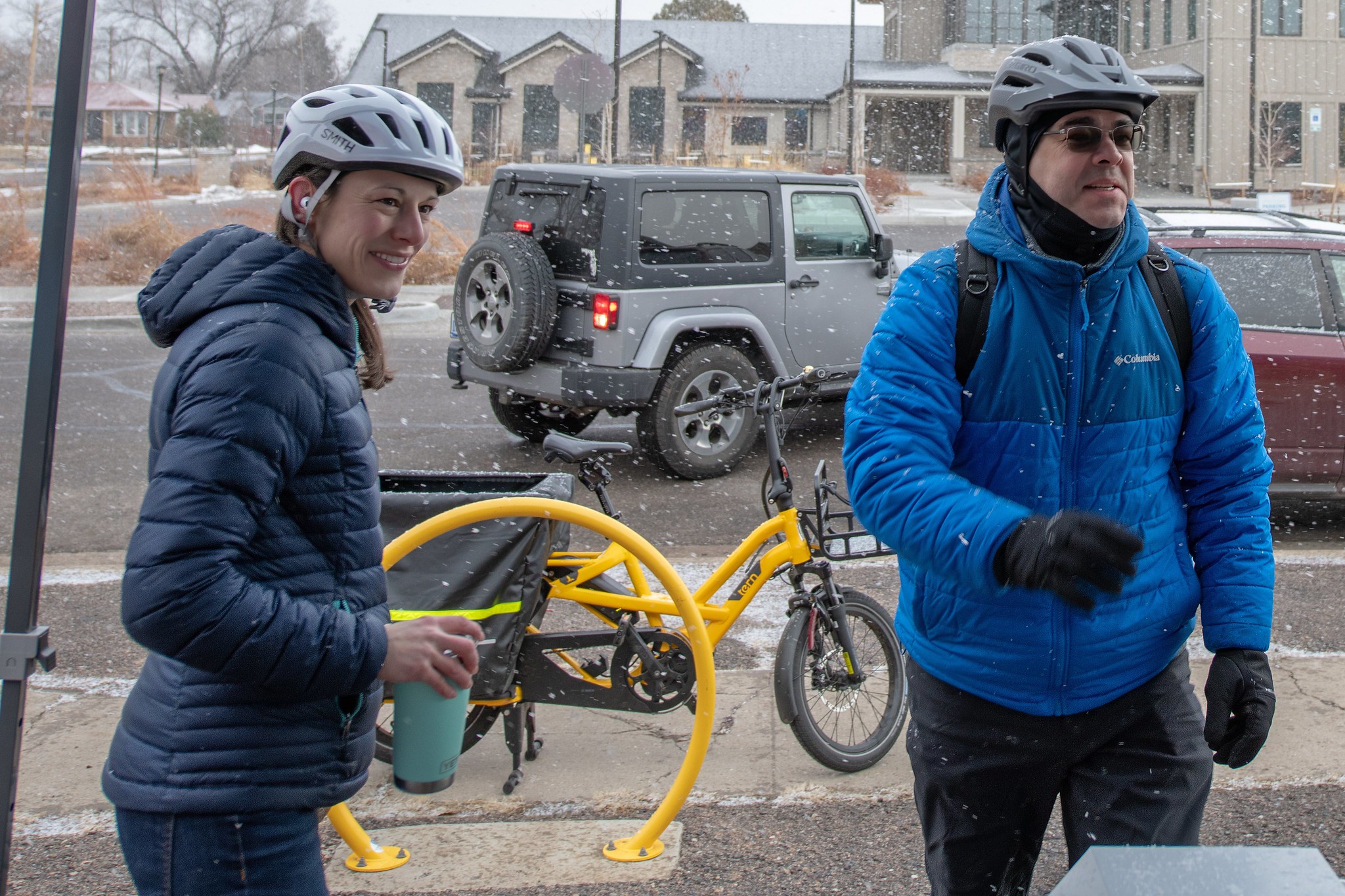 A man and woman stand outside in the snow with their bicycles.