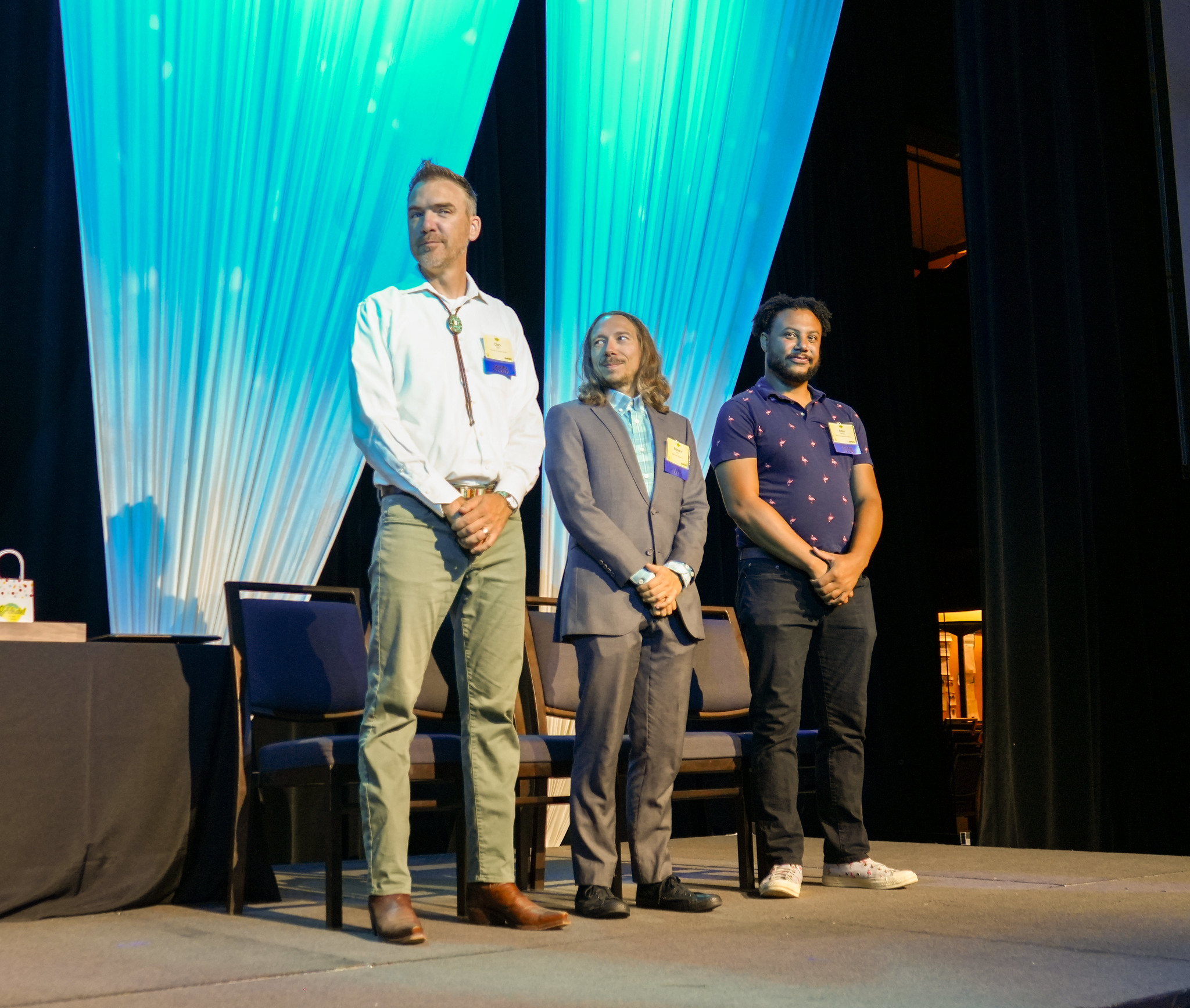 Three people stand on a stage with a blue curtain.