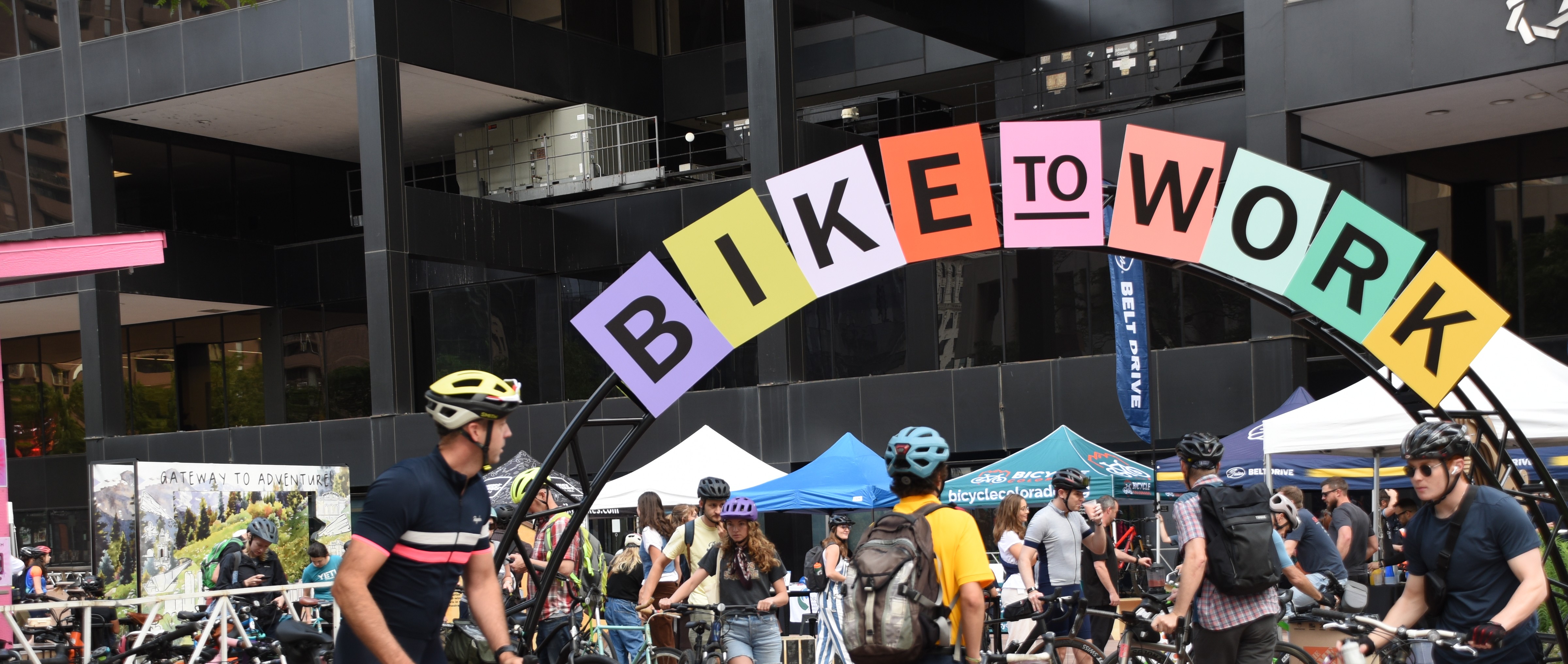 An arch with colored papers spelling out Bike to Work Day in front of an office building in downtown Denver.  A crowd of people wearing bike helmets and riding bikes congregate in front of outdoor tables.