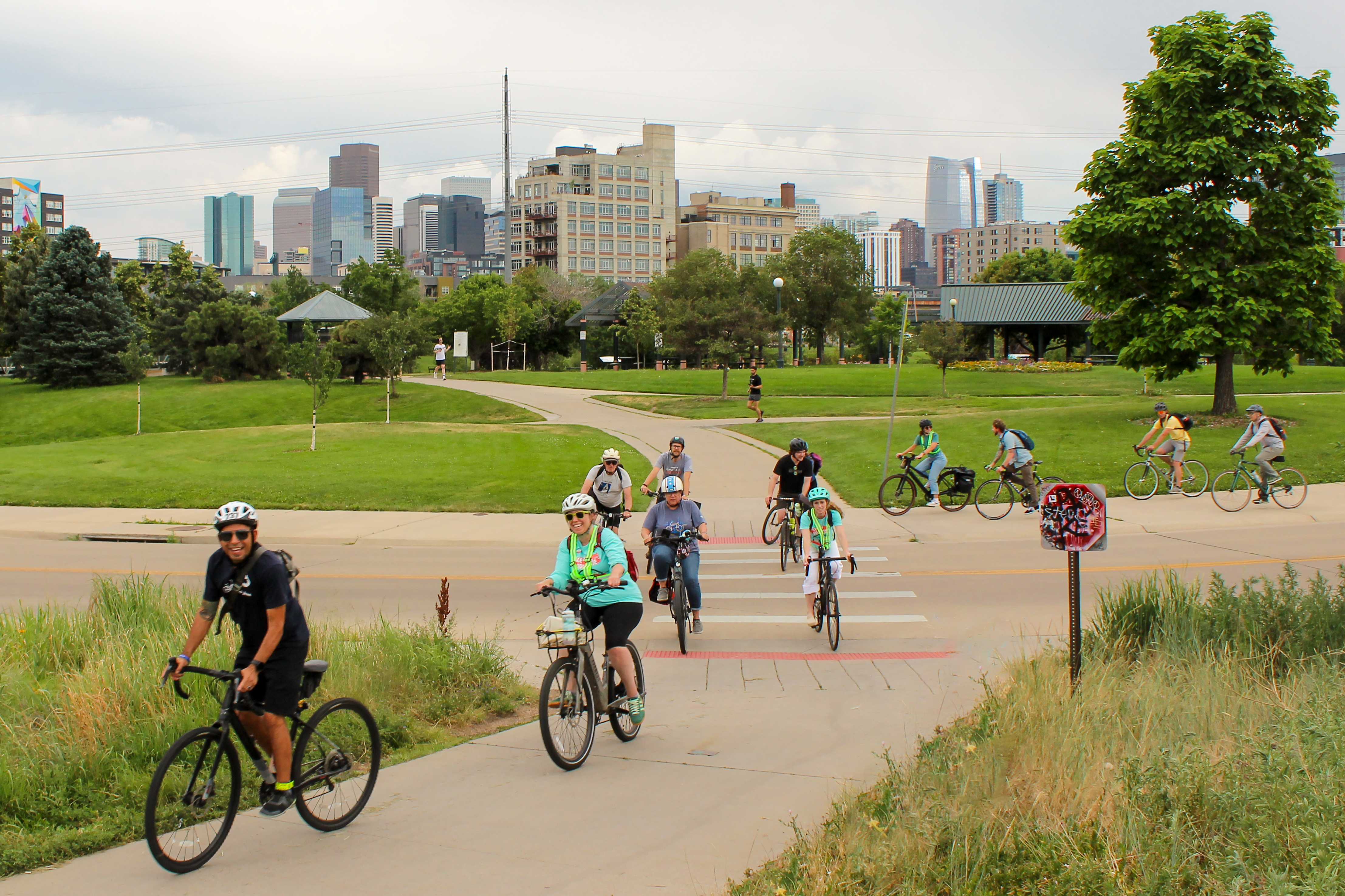 A group of cyclists cross a street intersection to reach a trail with downtown Denver in the background.
