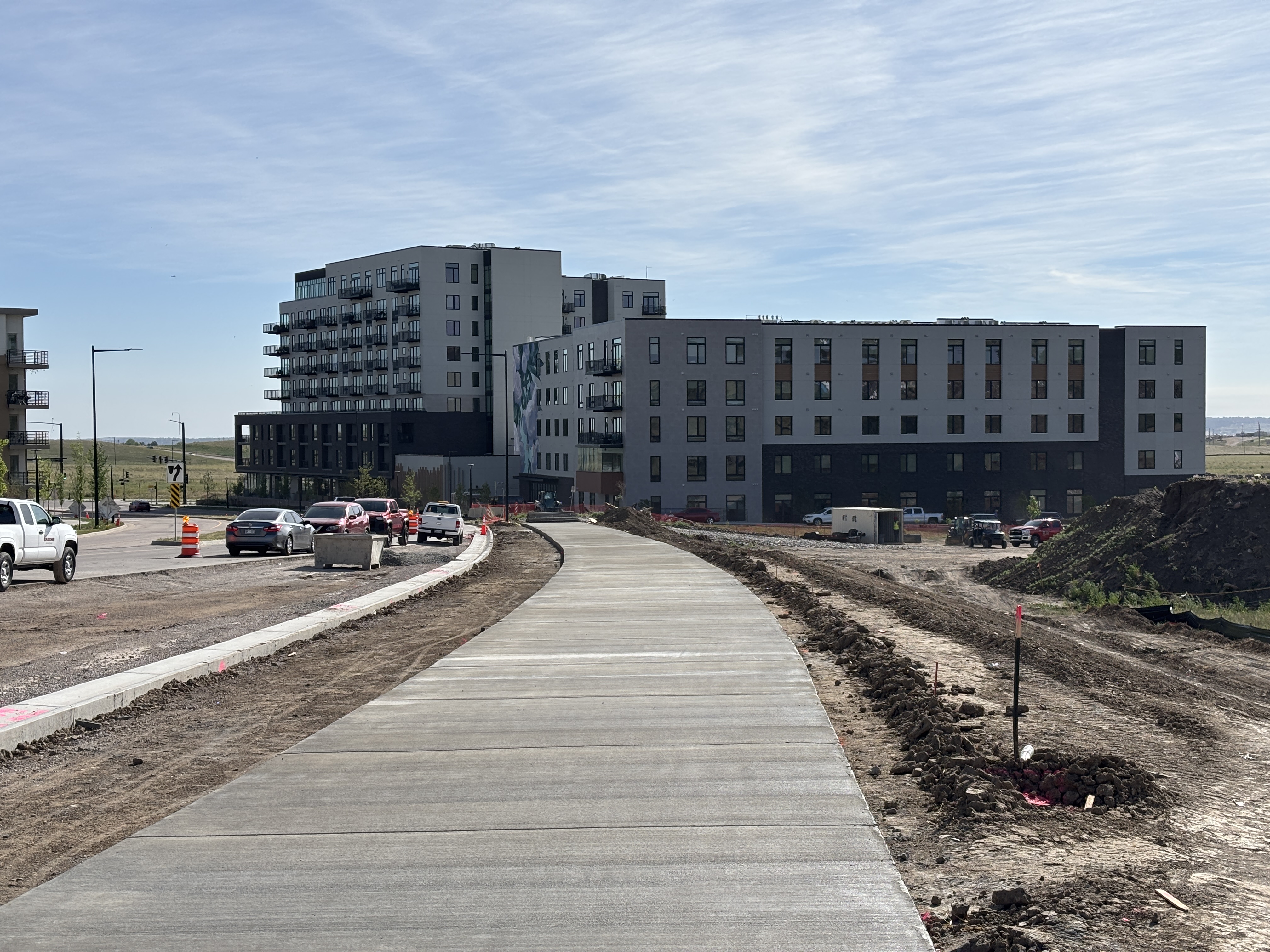 A new bike trail with dirt on either side runs parallel to a street, leading toward a modern high-rise residential building. 