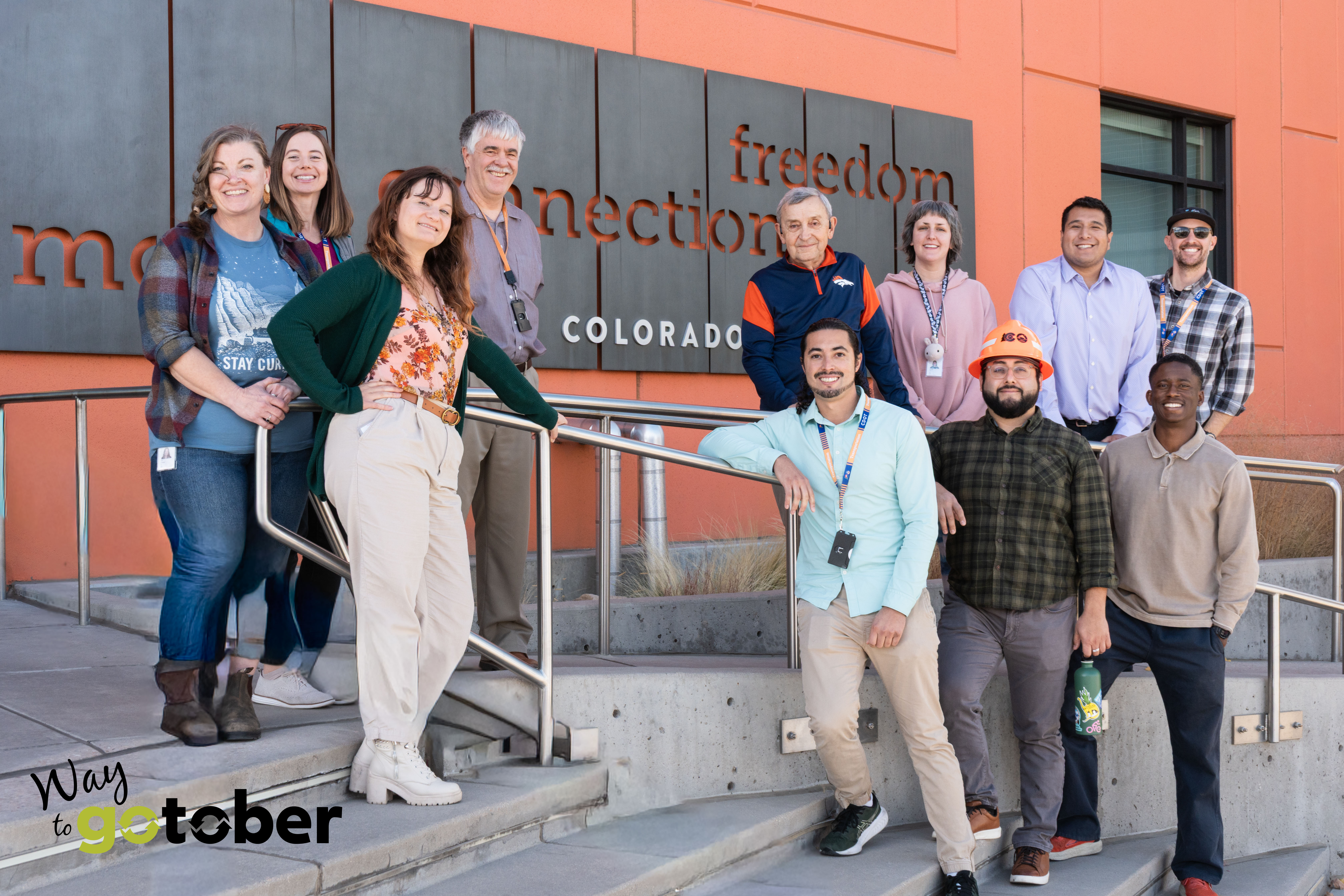 A group photo of Colorado Department of Transportation employees in front of an orange building leaning on a metal railing with the Way to Go-Tober logo in the bottom left corner.