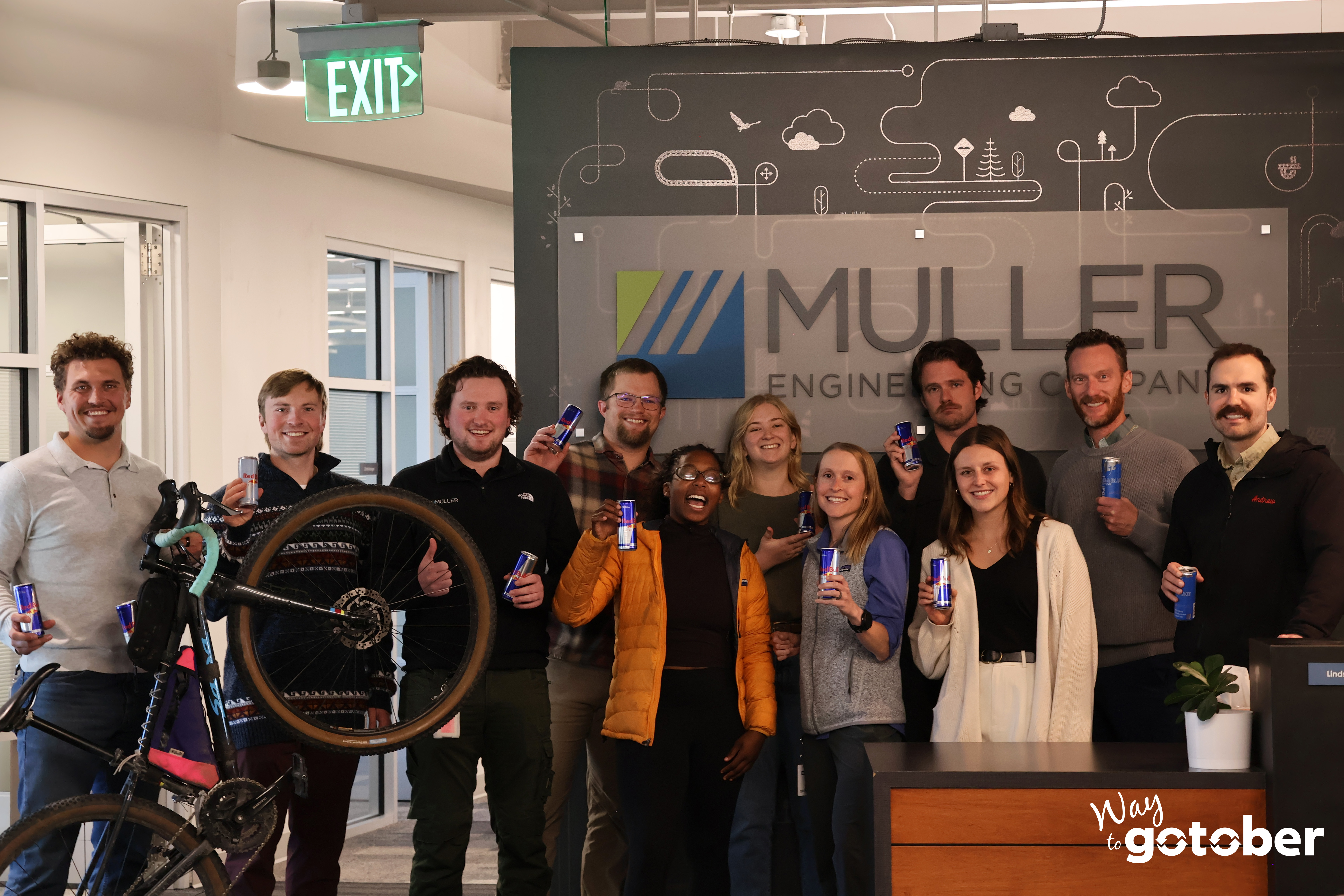 A group photo of Muller Engineering company employees in front of their company logo. Each individual is holding a can of Red Bull and a bicycle standing on it's bike wheel with the "Way to Go-tober" logo in the bottom right corner.