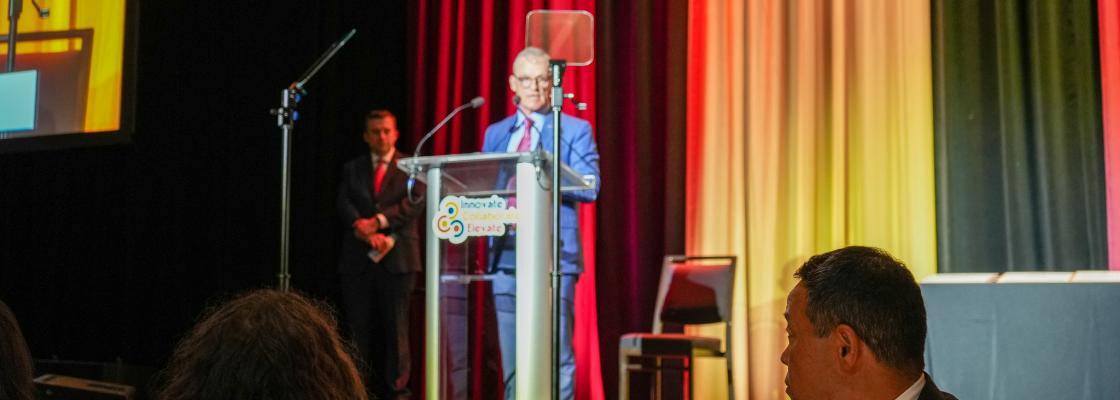 In a dimly lit ballroom, a middle-aged white man stands in front of a lectern giving remarks to 500 people.