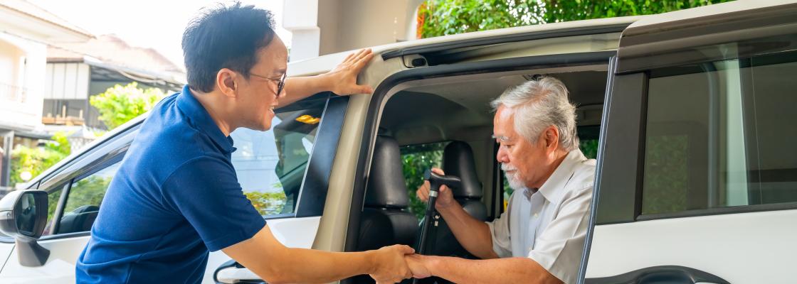 A man of Asian descent  helps an older adult man of Asian descent with a cane out of a white minivan.