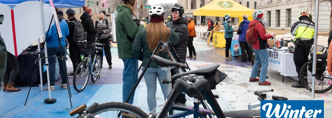 Cyclists gather with their bikes at an outdoor event with tents and information booths in a city plaza.