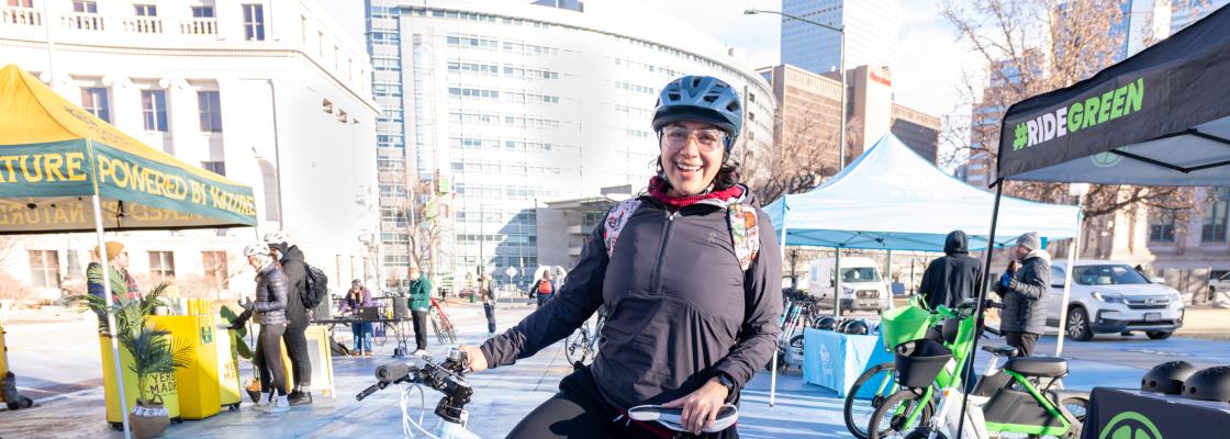 A cyclist stands with their white bike in a bright outdoor event space filled with tents, banners, and other riders.