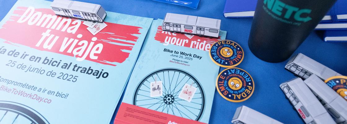 Bike to Work Day posters in English and Spanish sit on a table with Pedal Smile Repeat buttons and other swag items.