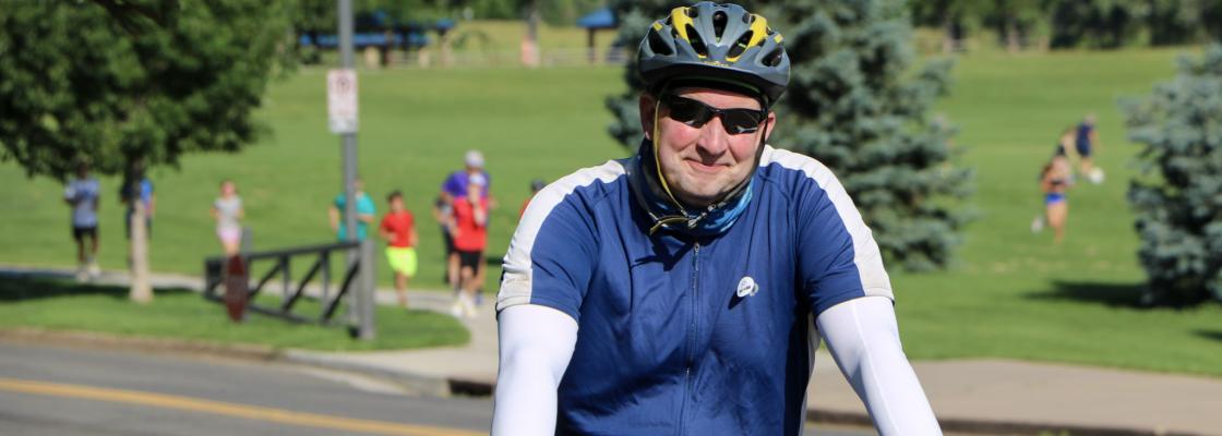 A male cyclist wearing a grey helmet, black sunglasses and a navy and white cycling kit smiles while seated on his bike with runners in a park featured in the background.