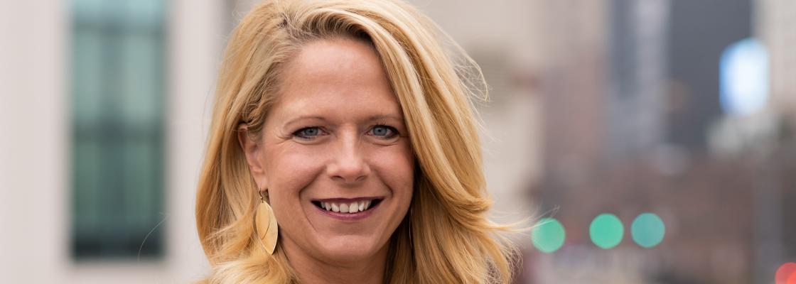 A middle-aged woman with long blond hair, wearing a black scoop-neck shirt, stands outdoors along a city street, smiling.