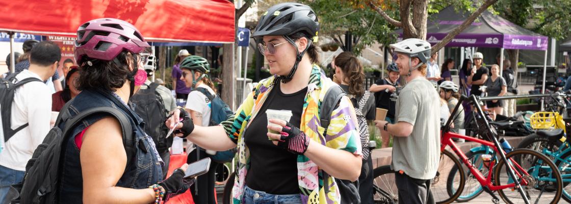 People with bicycles and helmets visit a Bike to Work Day commuter station, where riders gather, socialize and receive refreshments under event tents.