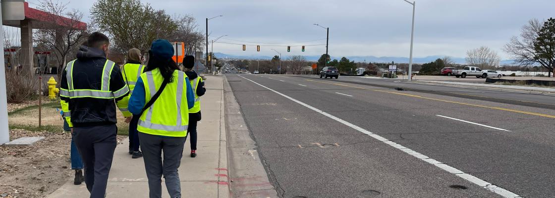 Several people wearing neon yellow safety vests walk down a sidewalk next to a four-lane road where a few cars are driving.